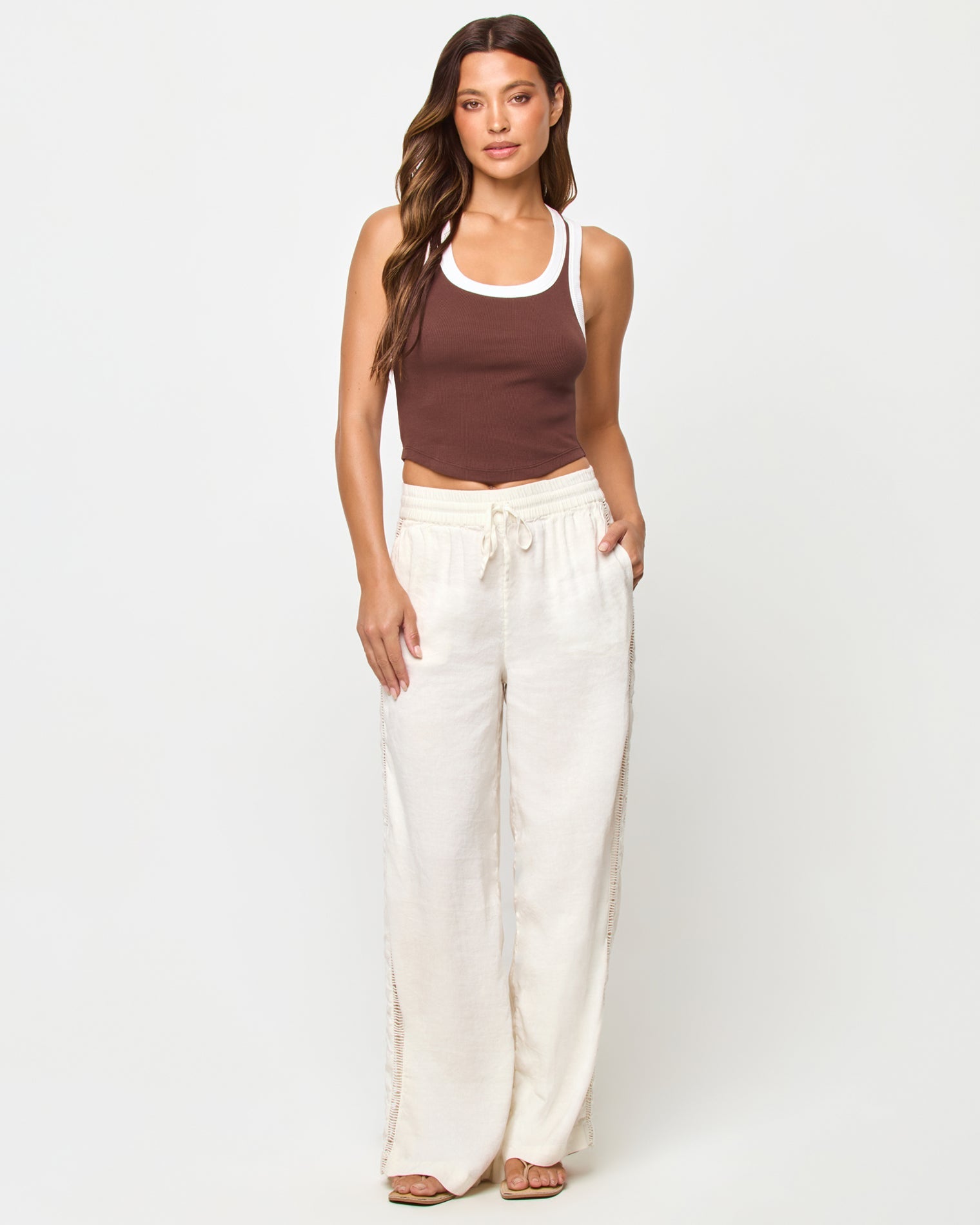 Full-length front view of a model wearing the LSPACE Ash Tank in espresso-cream, a cropped ribbed racerback tank with espresso brown body and cream contrast trim, styled with cream wide-leg drawstring pants and sandals.