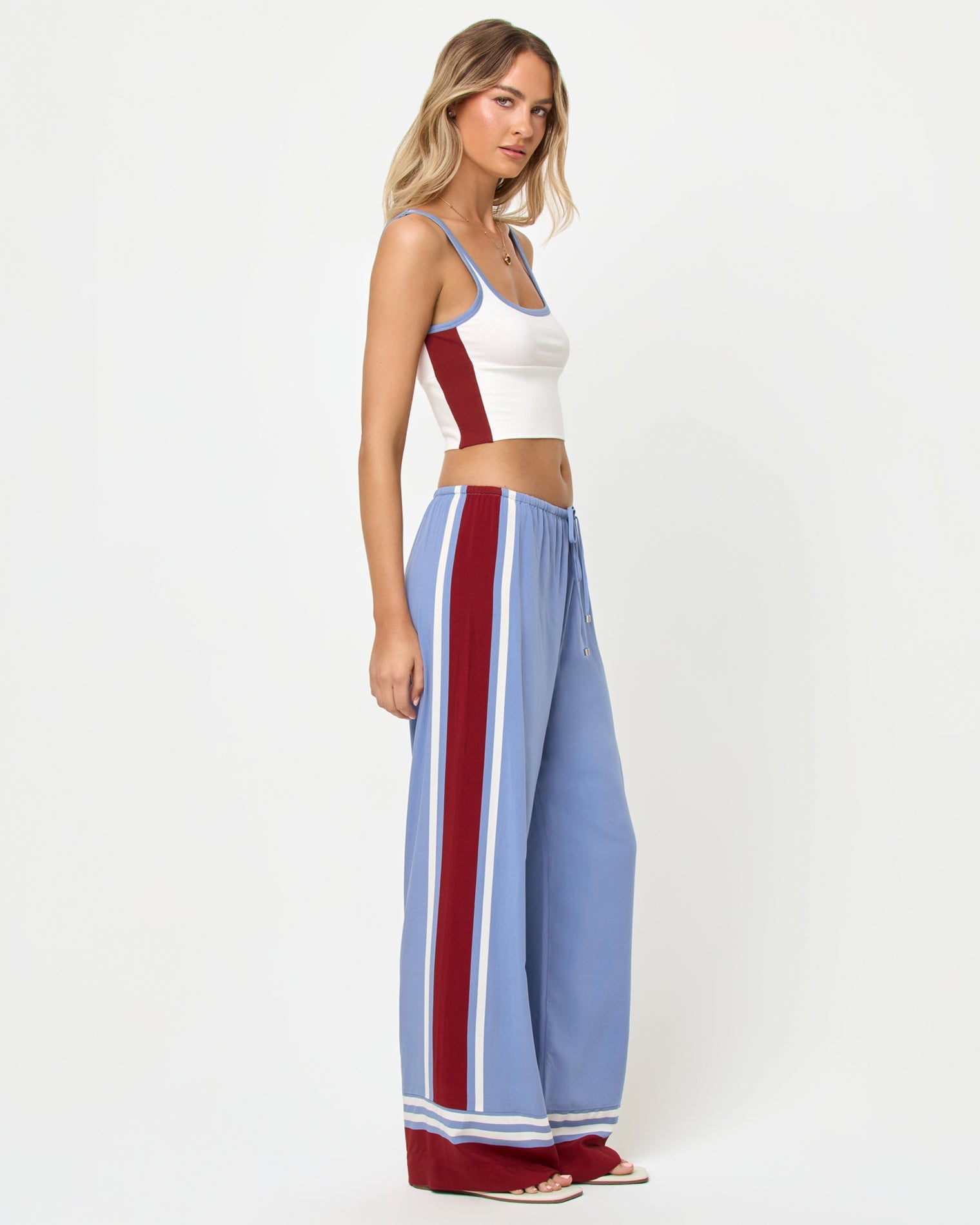 Three-quarter front side profile studio view of the Reset Tank and Unwind Pant in Cream-Chambray-Fig, a cream ribbed scoop-neck crop cami with chambray blue and burgundy trim and color-block sides paired with chambray blue wide-leg stripe pants with a wide burgundy vertical side stripe and cream piping, styled with nude heeled sandals.